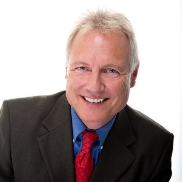 Chris Schueler in a black suit, blue shirt, and red tie. 