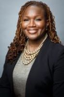 Andrea Anderson poses for a headshot in a black blazer, gray top, and gold necklace. 