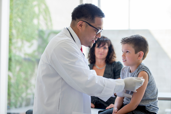 A little boy sitting on an examining table in a doctors office with his mother as the doctor gives him a shot.