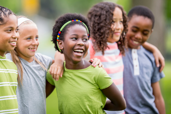 A group of elementary school kids are outside on a summer day. They are embracing and smiling while taking a group photo.