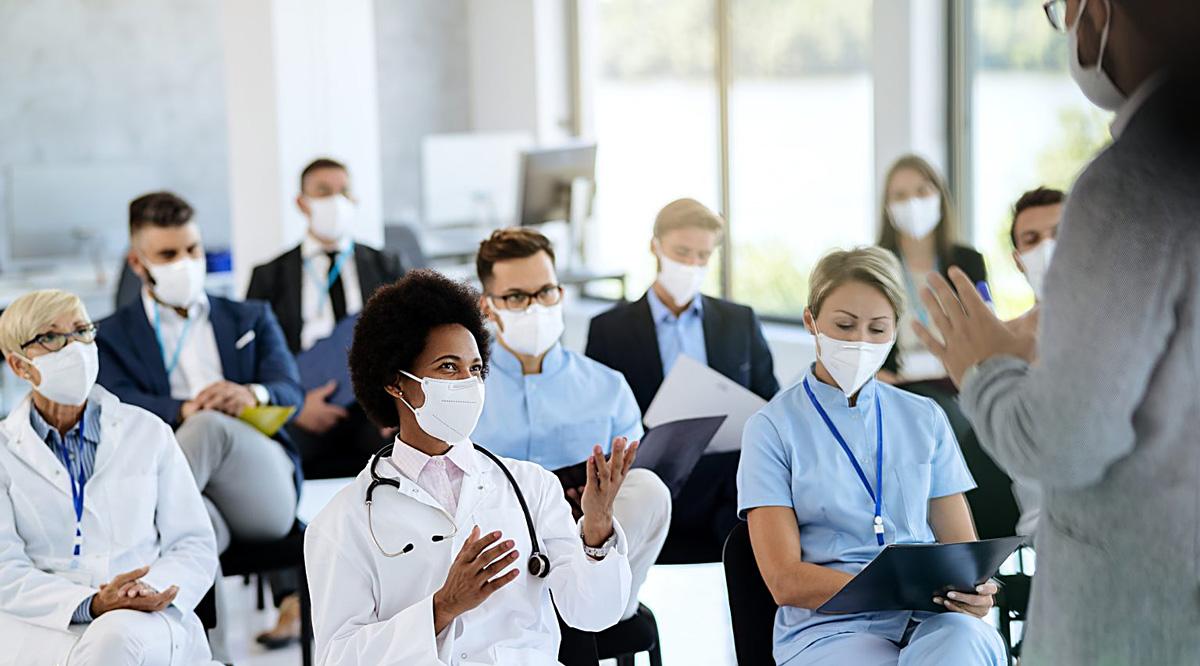 A healthcare worker talking to a presenter while attending a seminar during coronavirus epidemic. In the background is a diverse group of people wearing masks and sitting down.