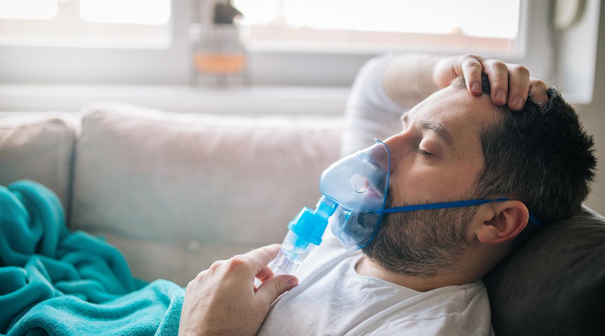 Mature adult man lying down on sofa in living room and using oxygen face mask.