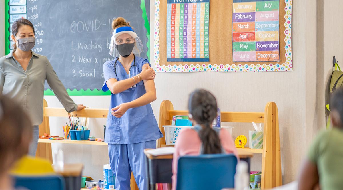 A school nurse standing at the front of a classroom speaking to a masked group of students about the Covid-19 vaccination.