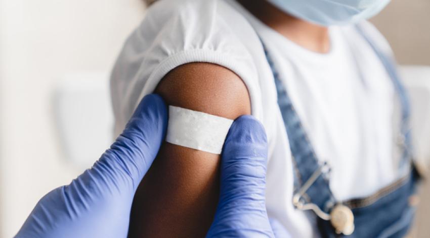 A gloved medical work places a band-aid on a masked child patient.