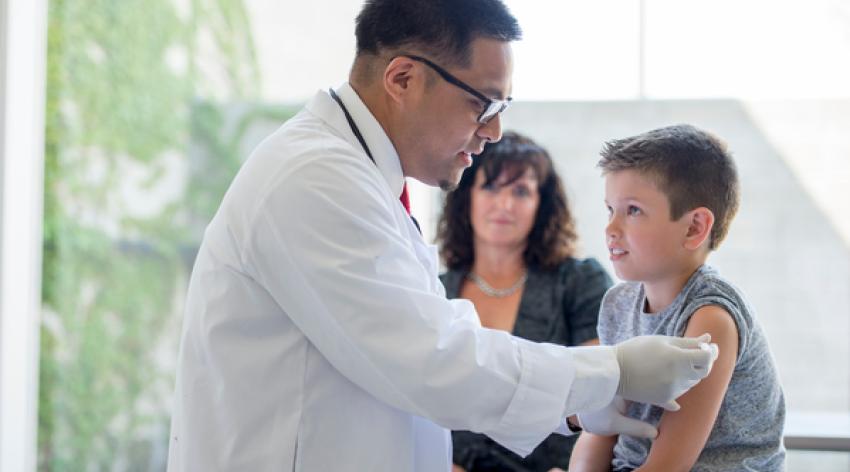 A little boy sitting on an examining table in a doctors office with his mother as the doctor gives him a shot.