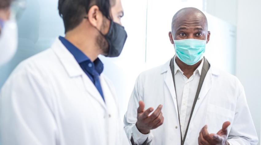 Two doctors wearing masks and white lab coats talking to each other. 
