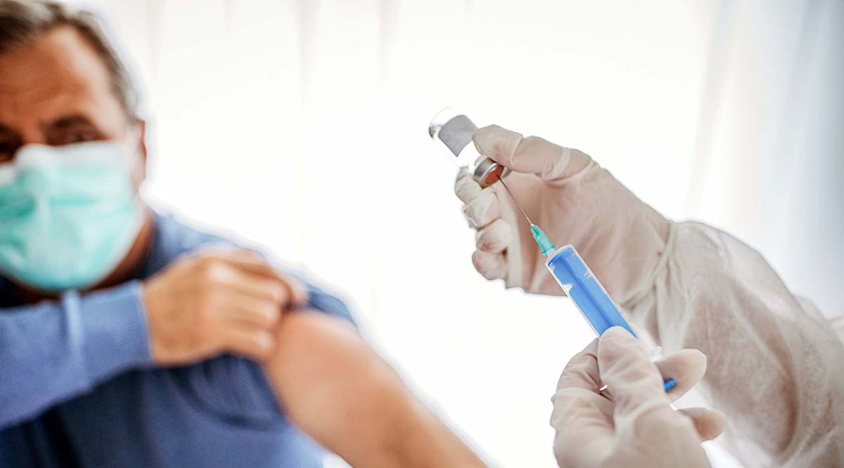 A masked patient pulls up his sleeve as a medical worker holds a syringe.